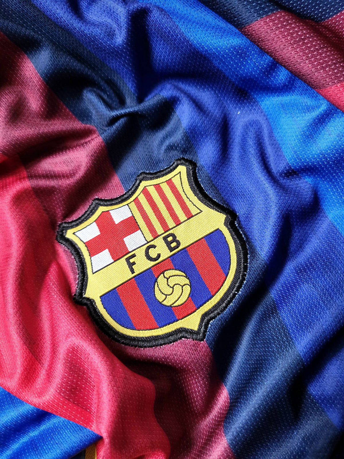 A close-up of an FC Barcelona soccer jersey fabric, showing the embroidered club crest on a textured blue and red striped mesh material.