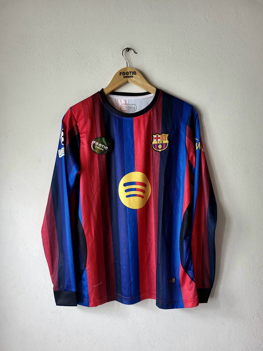 A long-sleeve FC Barcelona soccer jersey with red and blue vertical stripes, featuring the yellow Spotify sponsor logo on the chest, hanging on a wooden hanger against a plain light gray wall.