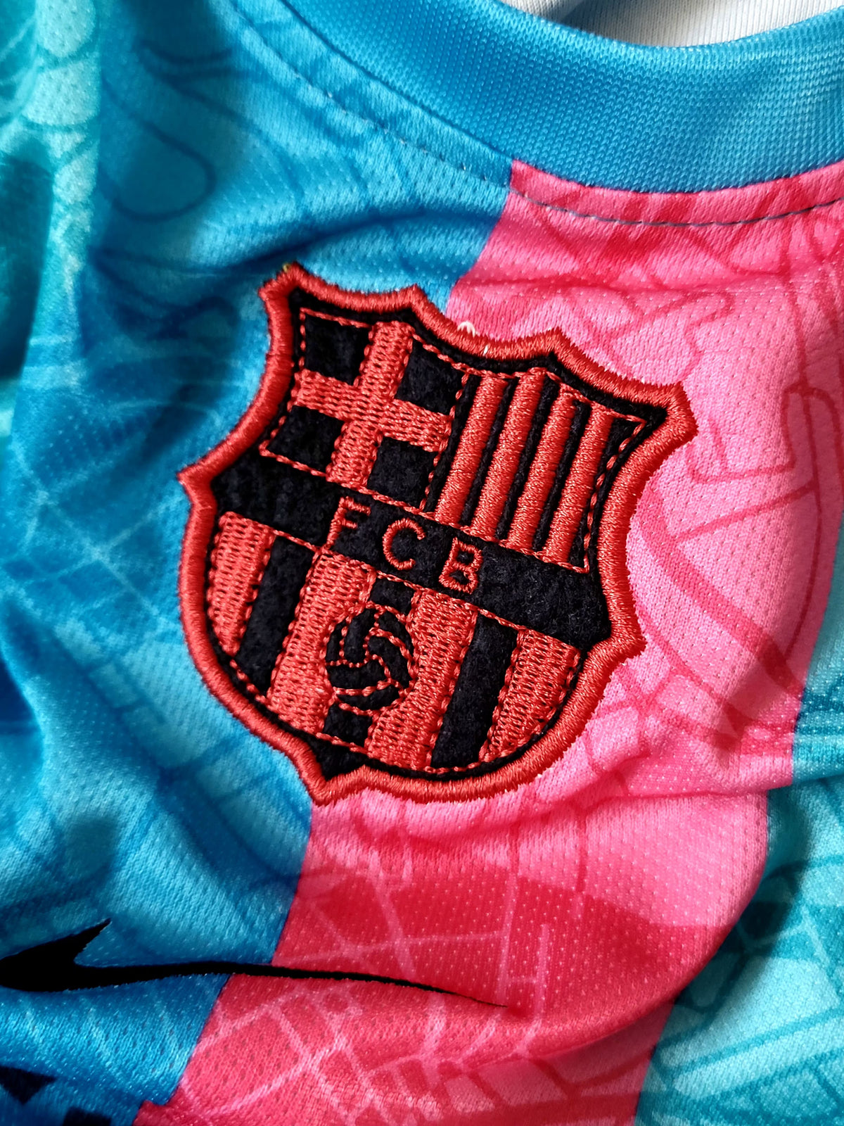 Close-up of an FC Barcelona soccer jersey showing the embroidered club crest in red and black over a split blue and pink patterned fabric background.