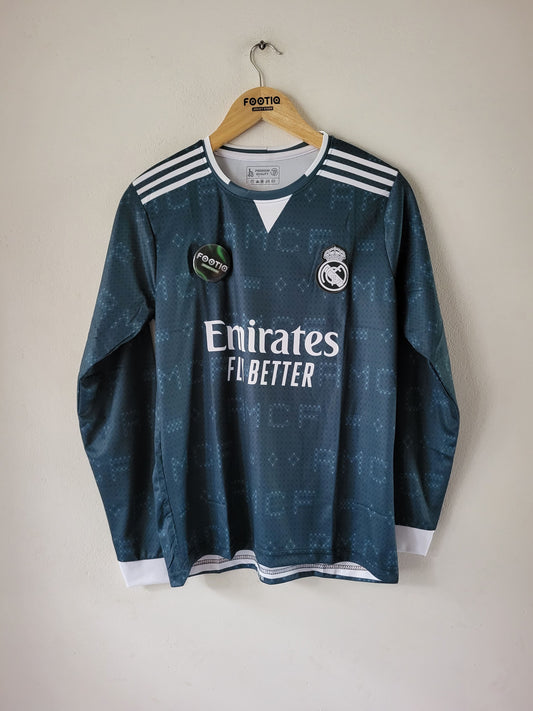 A dark teal Real Madrid soccer jersey with white accents, hanging on a wooden hanger against a plain beige wall. The jersey features the Emirates Fly Better sponsor logo, the Real Madrid crest, and Adidas shoulder stripes.
