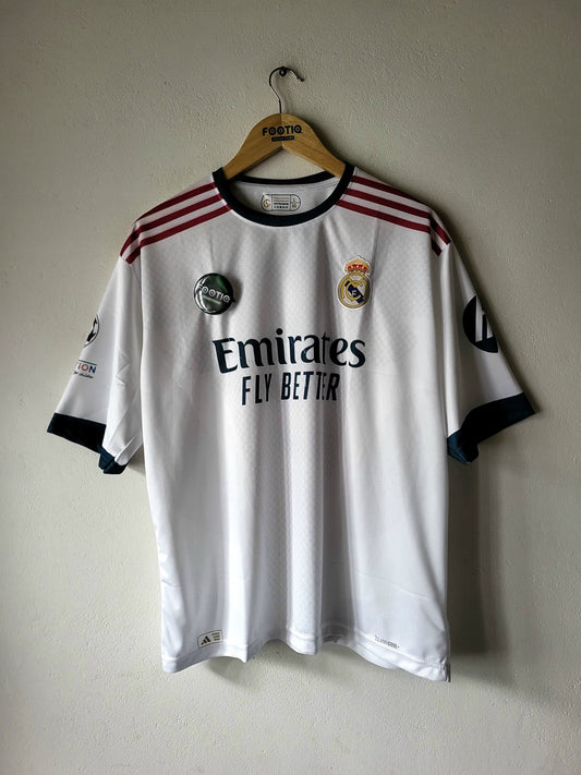A white Real Madrid soccer jersey with 'Emirates Fly Better' sponsor text, hanging on a wooden hanger against a plain wall.