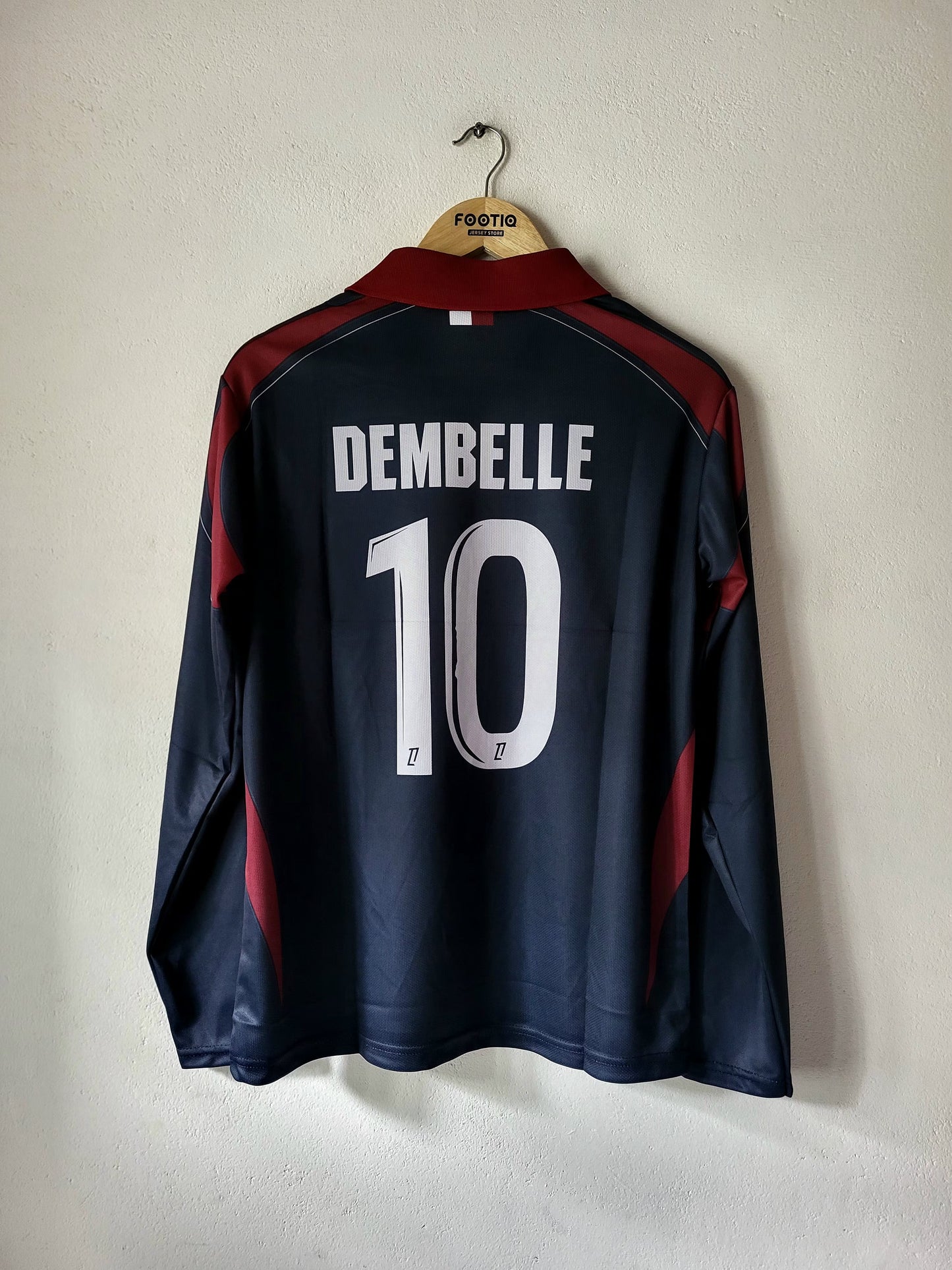 A dark navy blue long-sleeve soccer jersey with 'DEMBELLE' and the number '10' printed in white on the back, hanging on a wooden hanger against a plain off-white wall.
