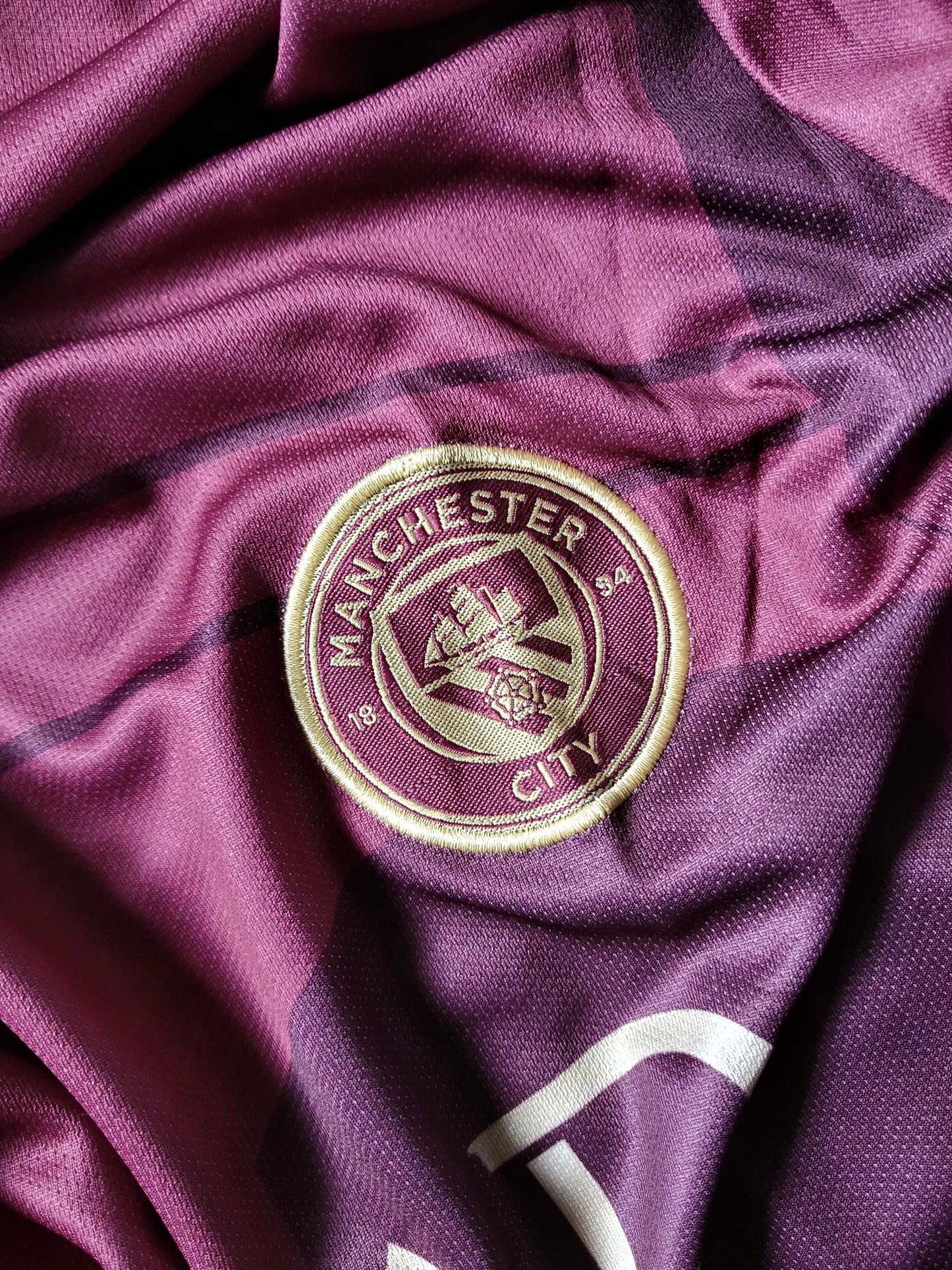 A close-up of a Manchester City soccer jersey in a deep purple color, featuring the embroidered club crest in gold and white on the chest.
