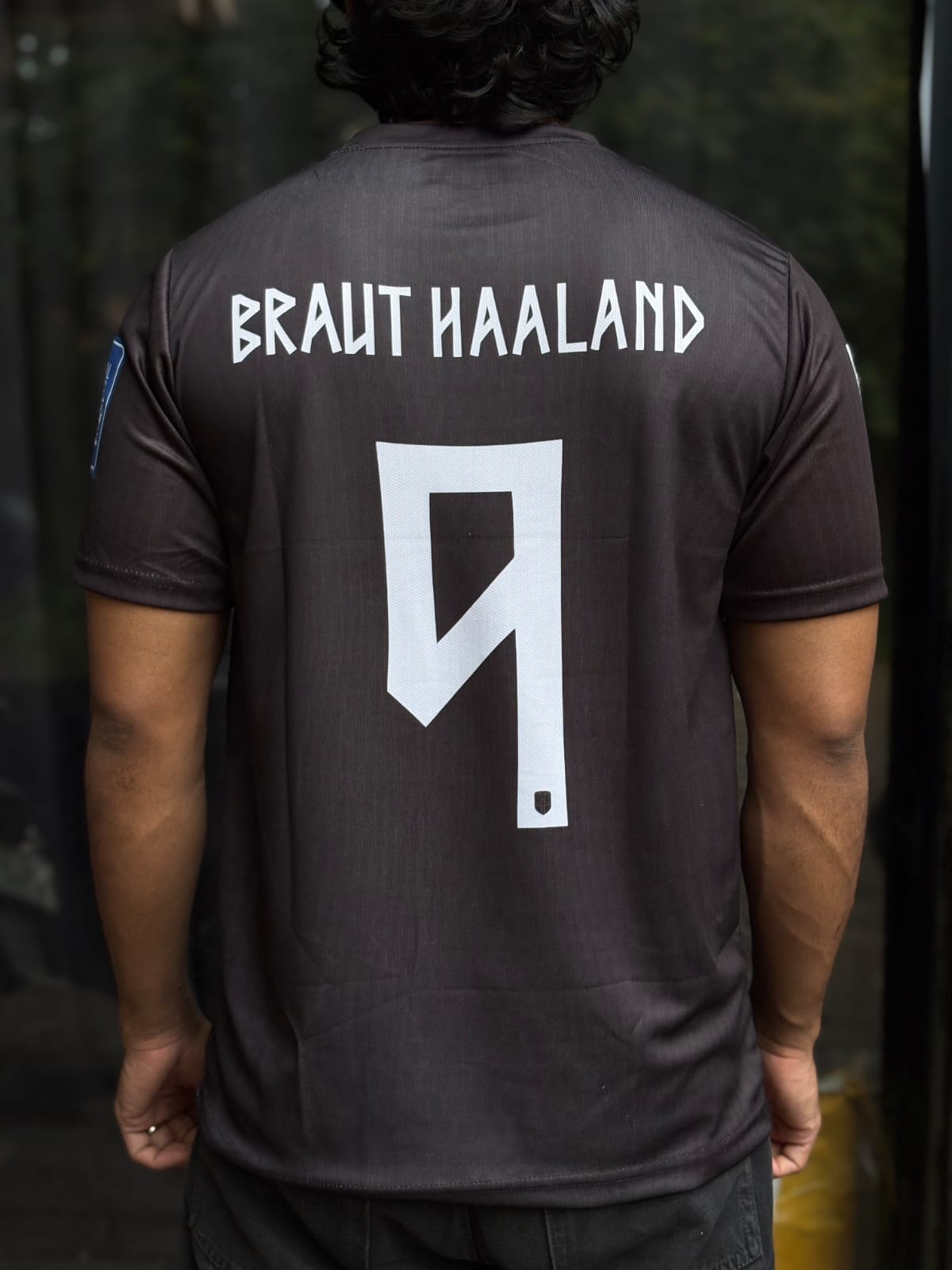Back view of a person wearing a dark brown short-sleeve soccer jersey with 'BRAUT HAALAND' and the number 9 printed in white on the back.