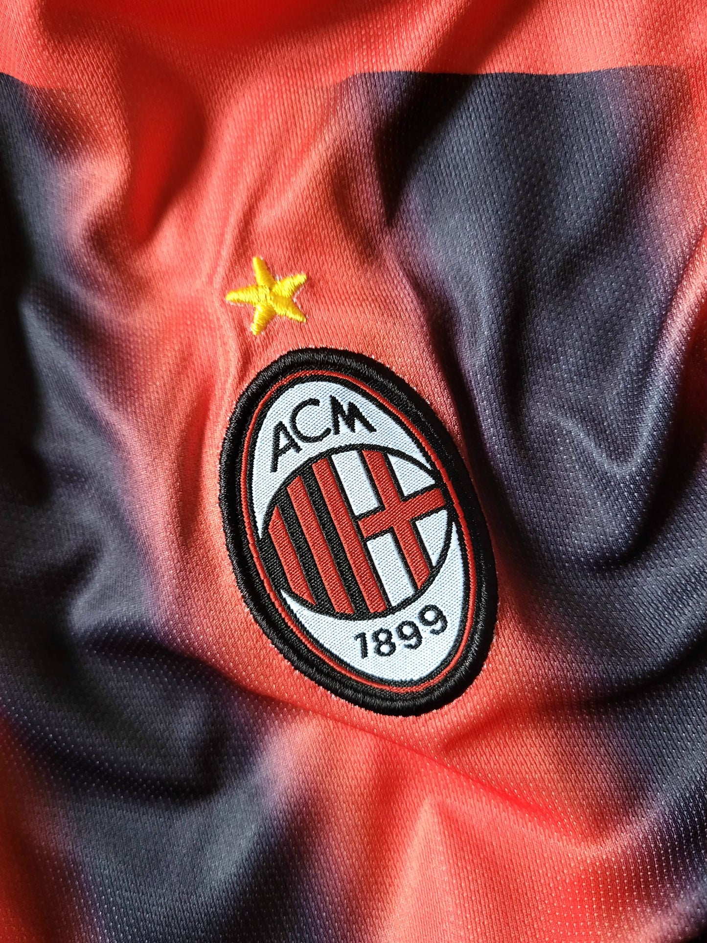 Close-up of an AC Milan soccer jersey fabric showing the embroidered club crest with red, black, white, and yellow details, plus a yellow star on a red-to-black gradient background.