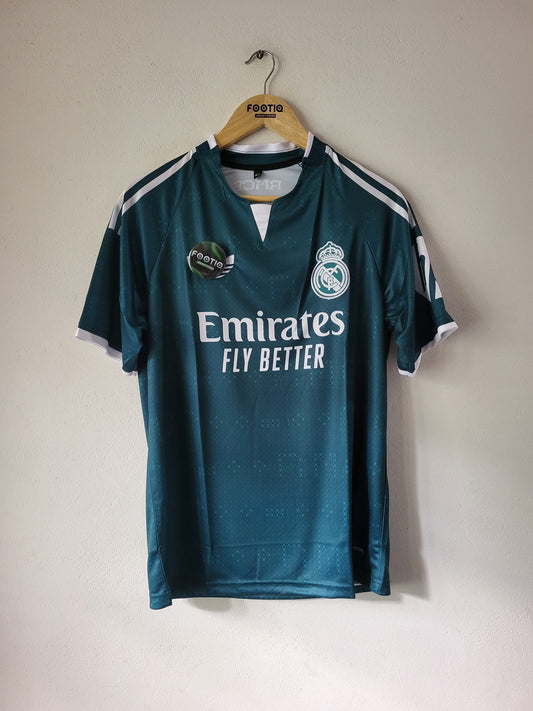 A dark teal Real Madrid soccer jersey hanging on a wooden hanger against a plain off-white wall. The jersey features white 'Emirates FLY BETTER' text on the chest, the Real Madrid crest, and shoulder stripes.