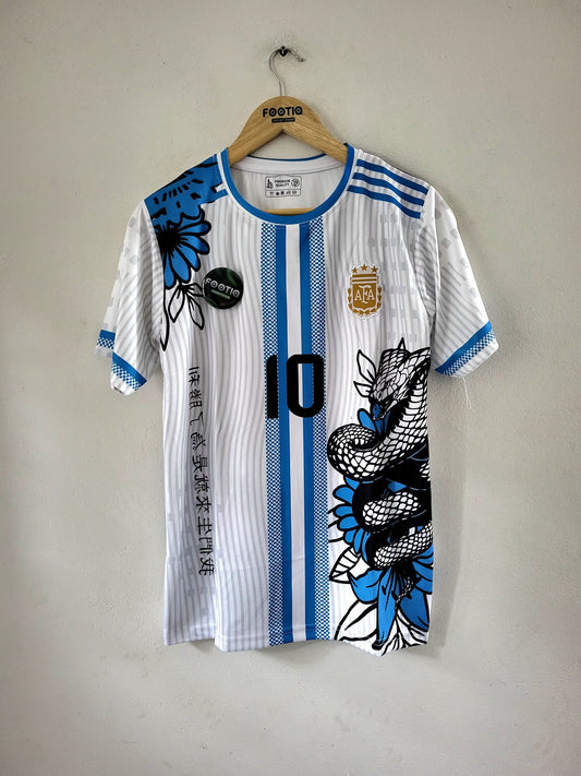 A white and blue striped Argentina soccer jersey with black and blue dragon and floral graphics, hanging on a wooden hanger against a plain wall.