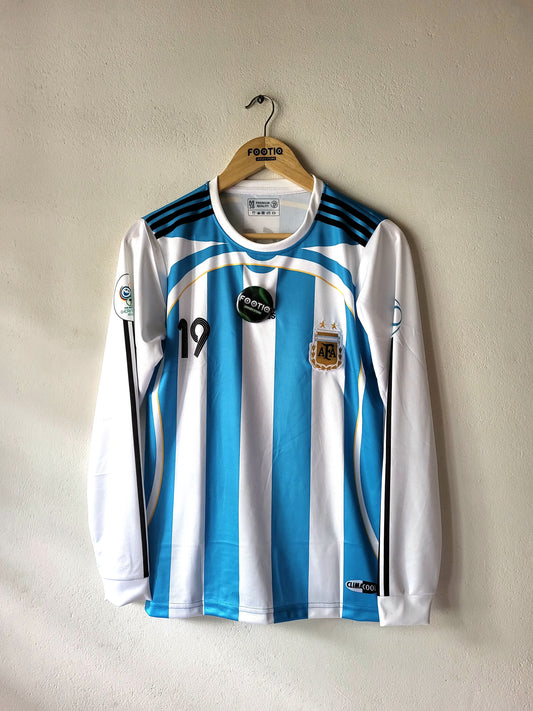 An Argentina national football team long-sleeve jersey with light blue and white vertical stripes, hanging on a wooden hanger against a textured beige wall. The jersey features black shoulder stripes, the number 19 on the right chest, and the AFA crest on the left chest.