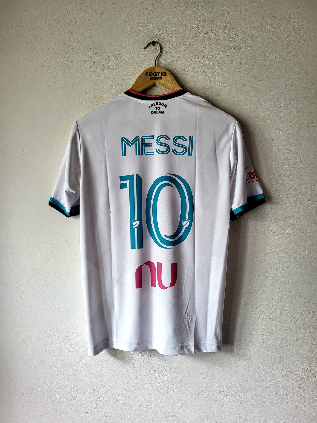 A white FC Barcelona soccer jersey with 'MESSI' and the number '10' printed on the back in blue, hanging on a wooden hanger against a textured beige wall.