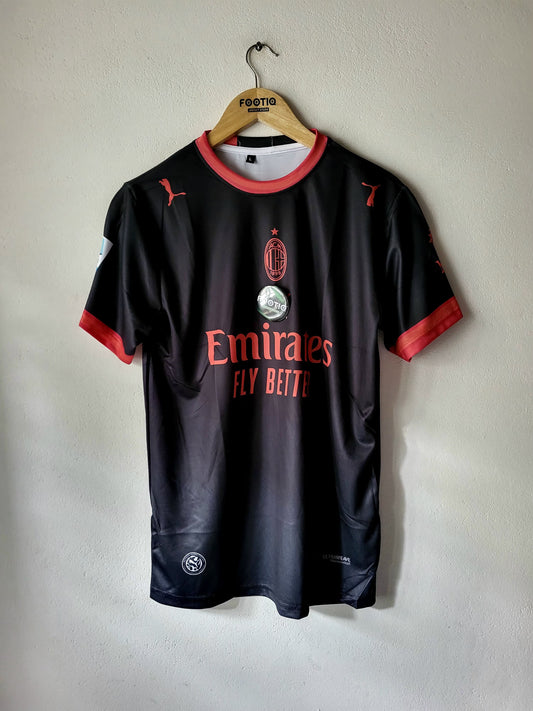 A black AC Milan soccer jersey with red trim, hanging on a wooden hanger against a light gray wall. The jersey features the AC Milan crest, the Emirates Fly Beite sponsor logo, and Puma branding.