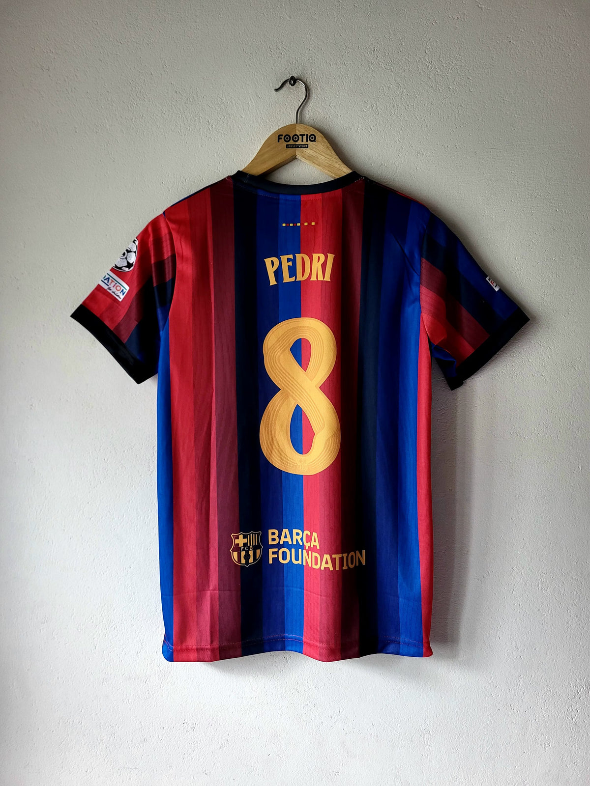 FC Barcelona home jersey with the name PEDRI and number 8 on the back, hanging on a wooden hanger against a light gray wall.
