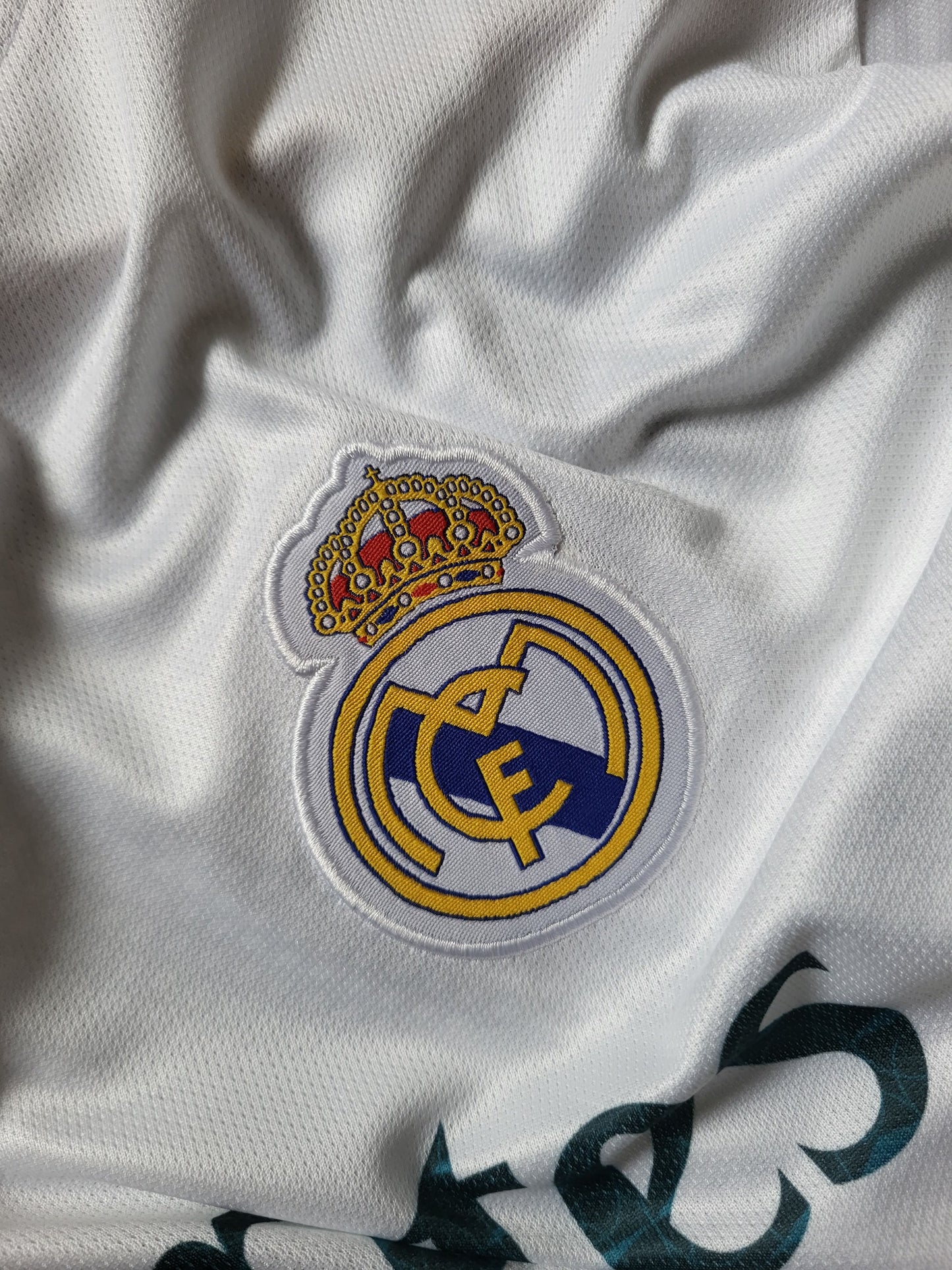 Close-up of a white Real Madrid soccer jersey fabric showing the embroidered club crest and a small gold-and-red crown patch on the chest.