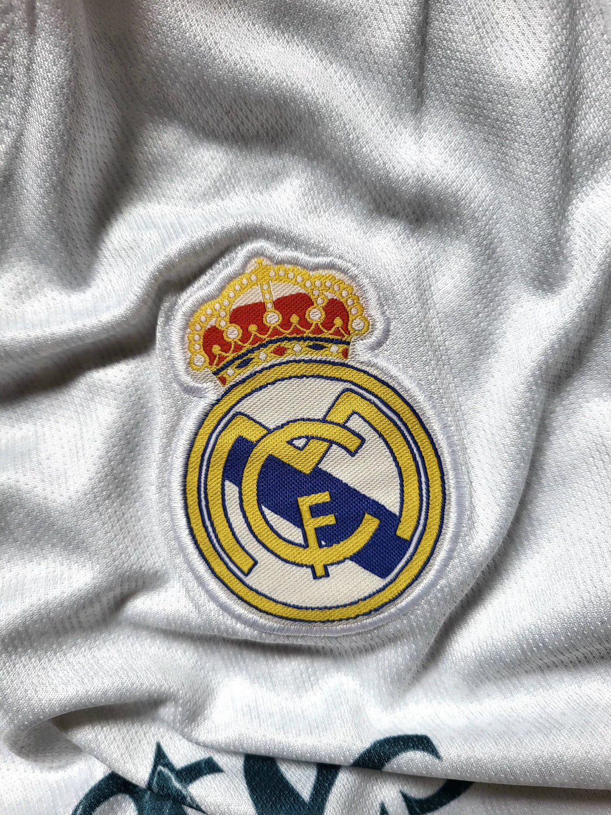 Close-up of a white Real Madrid soccer jersey showing the embroidered club crest with a gold crown on the chest.