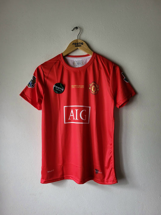 A red Manchester United short-sleeve soccer jersey hanging on a wooden hanger against a plain light gray wall. The jersey features the AIG sponsor logo on the front, the Manchester United crest, and various patch badges on the sleeves and chest.