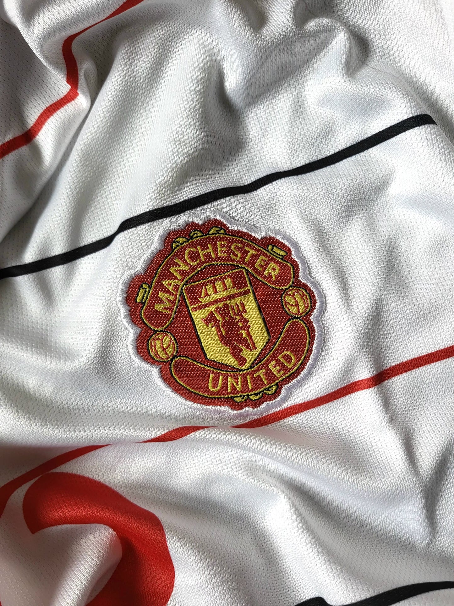 Close-up of a Manchester United football jersey fabric showing the embroidered club crest on a white textured surface with red and black striping.