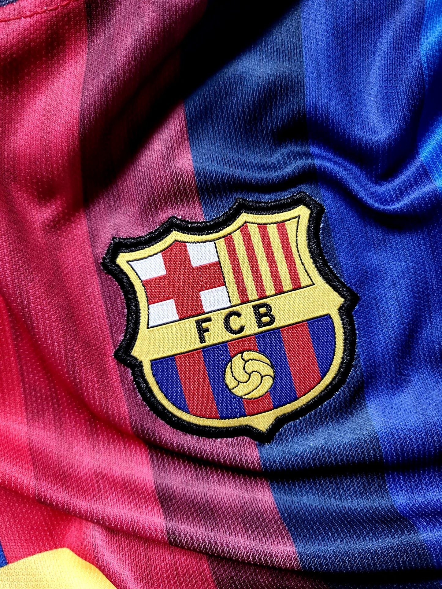 Close-up of an FC Barcelona embroidered crest patch on a satin-textured red and blue striped soccer jersey.