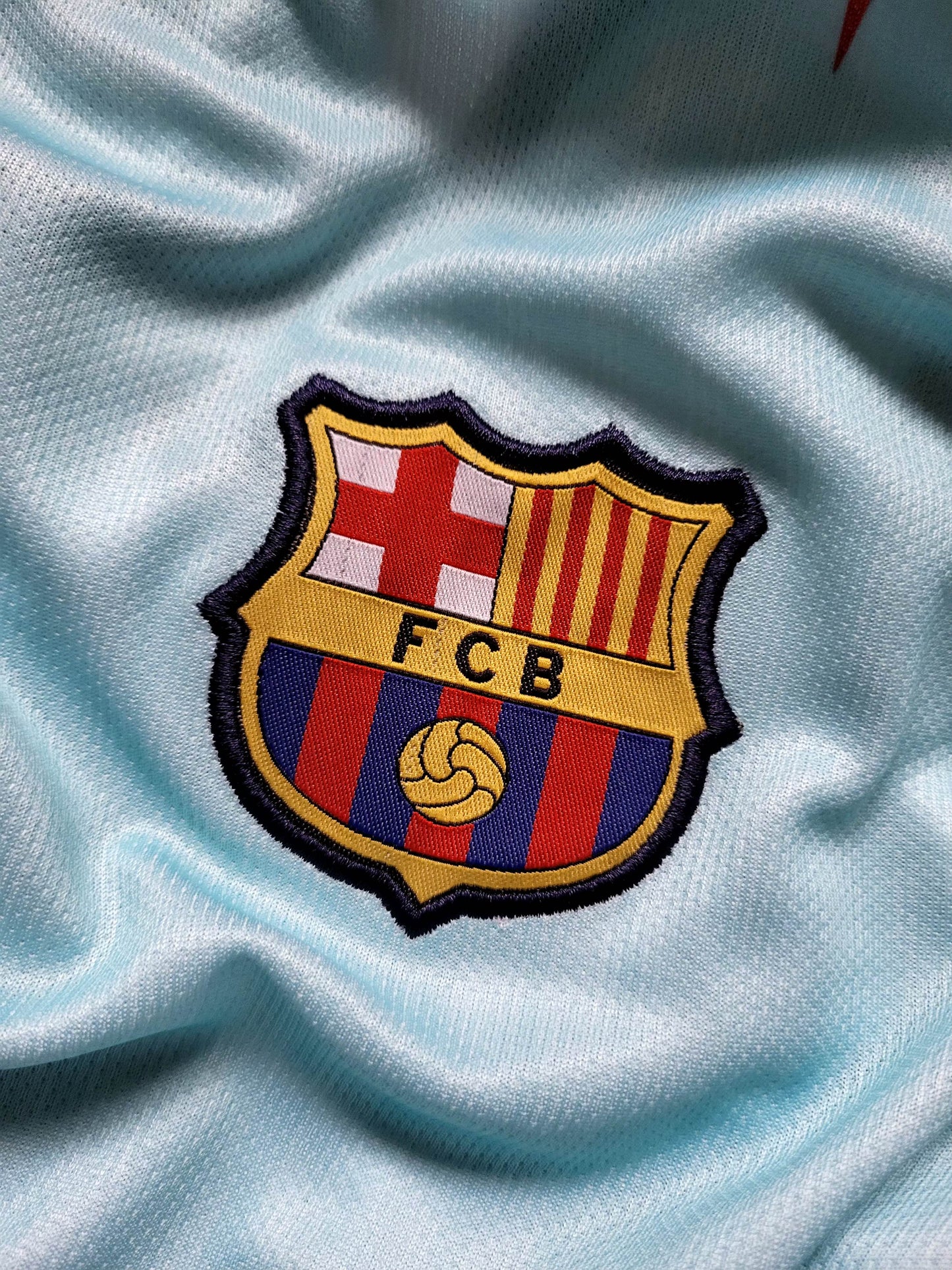 A close-up of the embroidered FC Barcelona crest patch on a light blue athletic jersey fabric.