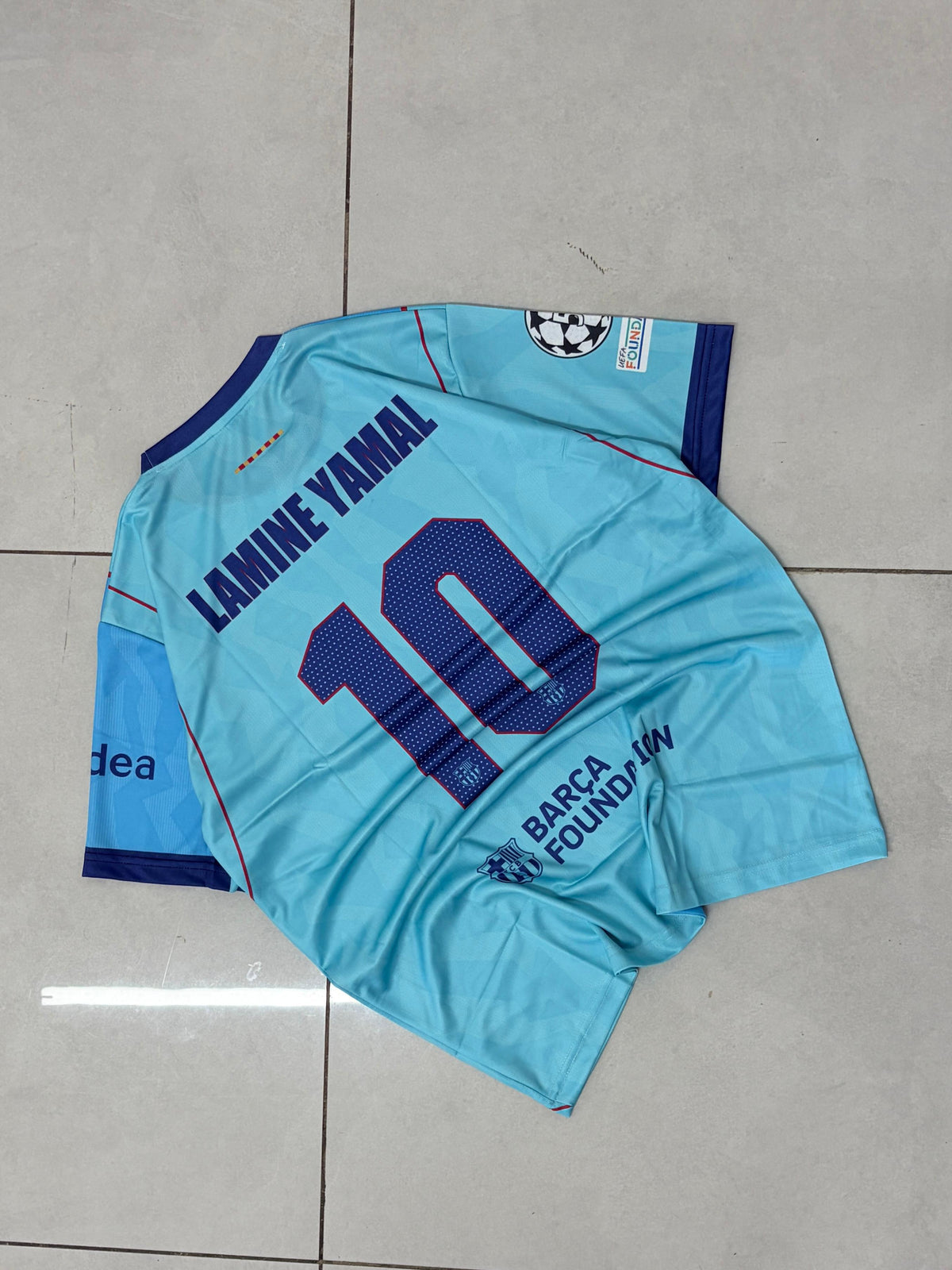 A light blue FC Barcelona soccer jersey with 'LAMINE YAMAL 10' printed on the back, laid flat on a gray tiled floor.