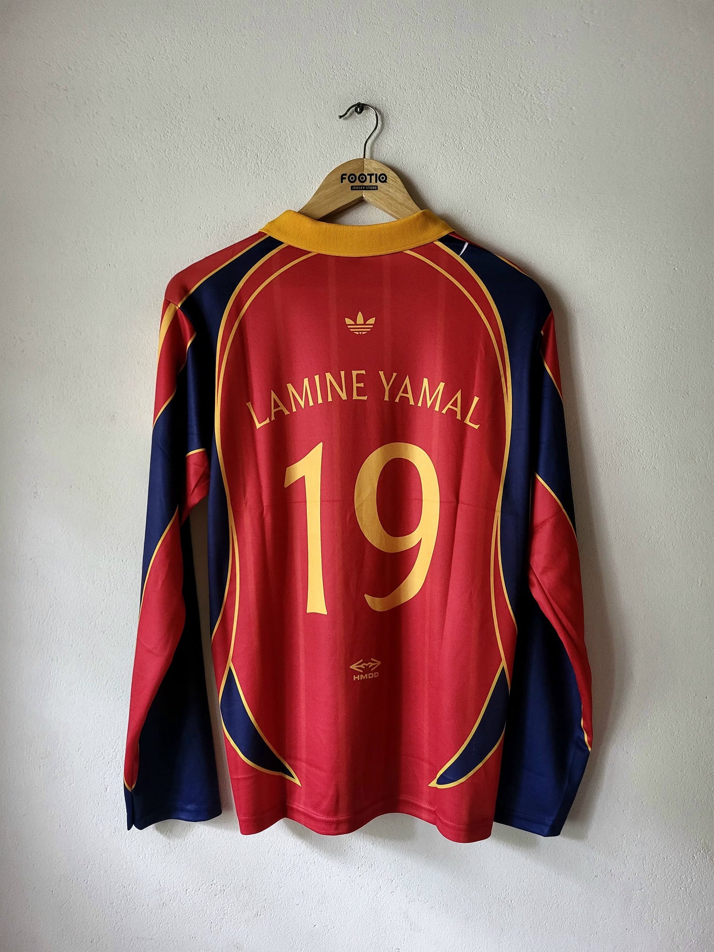 A red and navy blue FC Barcelona long-sleeve soccer jersey with 'LAMINE YAMAL' and the number '19' printed in gold on the back, hanging on a wooden hanger against a plain wall.