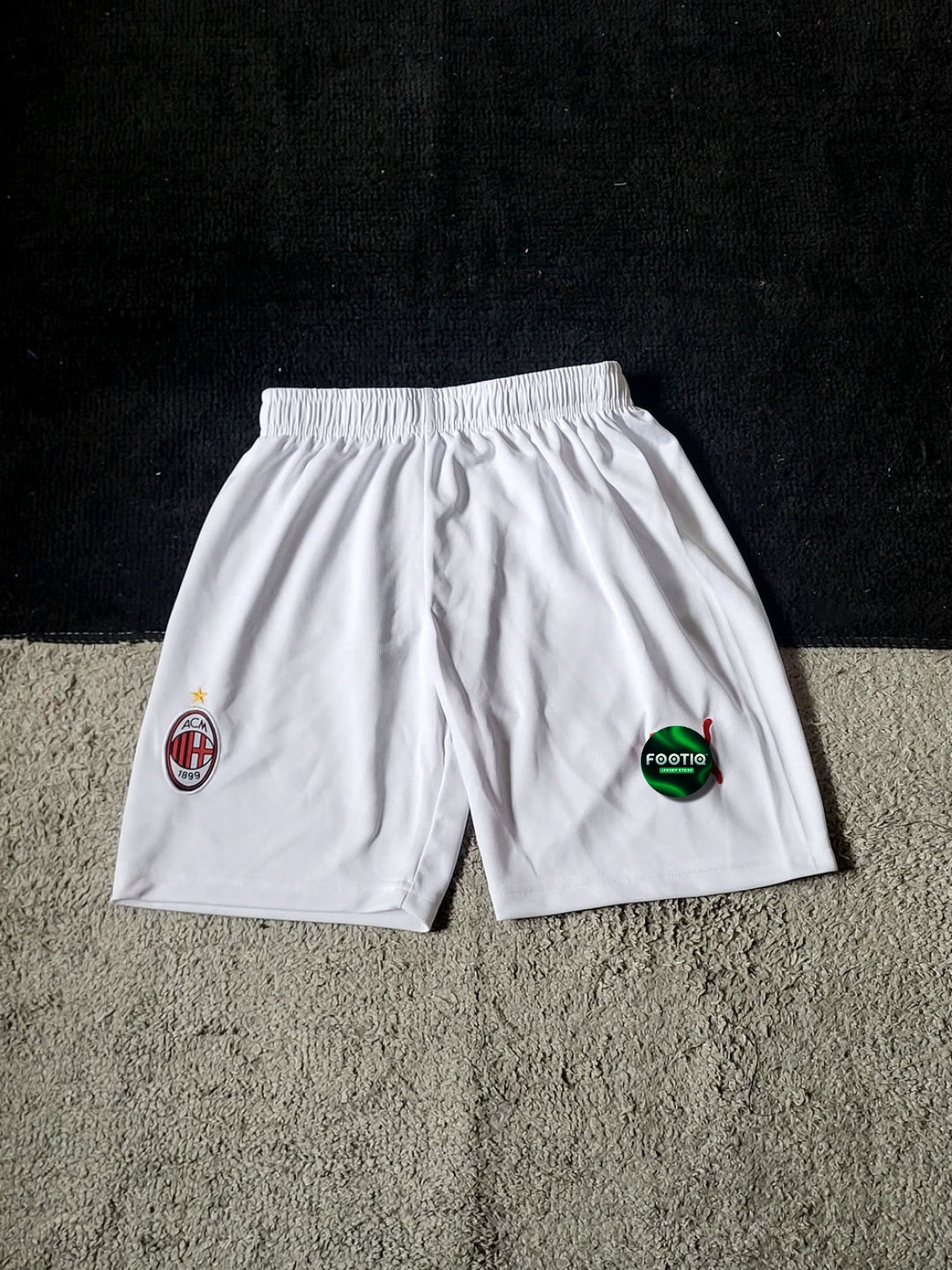 White AC Milan soccer shorts with the club's crest on the left leg and a green sponsor logo on the right leg, laid flat on a textured gray carpet against a black background.