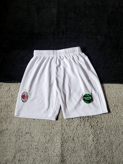 White AC Milan soccer shorts with the club's crest on the left leg and a green sponsor logo on the right leg, laid flat on a textured gray carpet against a black background.
