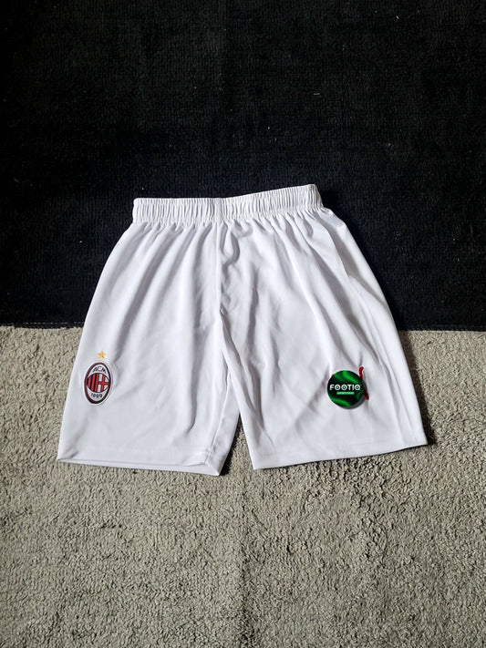 White AC Milan soccer shorts with the club's crest on the left leg and a green sponsor logo on the right leg, laid flat on a textured gray carpet against a black background.