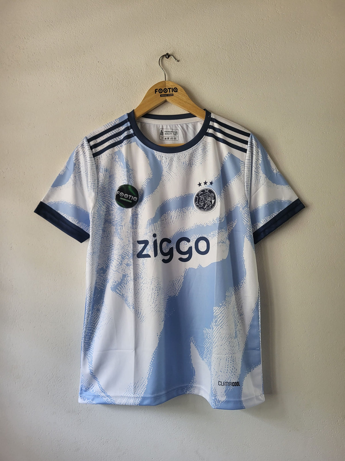 A short-sleeve Ajax football jersey in a white and light blue abstract pattern, hanging on a wooden hanger against a beige wall. The jersey features navy blue trim, three stripes on the shoulders, the Ajax club crest, the 'ziggo' sponsor logo, and the Pinnacle logo.