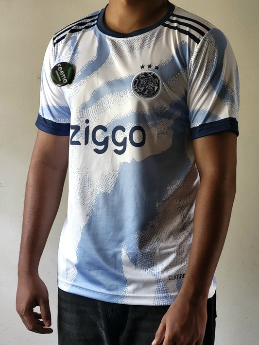 A person wearing a short-sleeve Ajax football jersey with a white and light blue abstract pattern, navy collar and sleeve trim, the Ziggo sponsor logo on the front, and the Ajax club crest.
