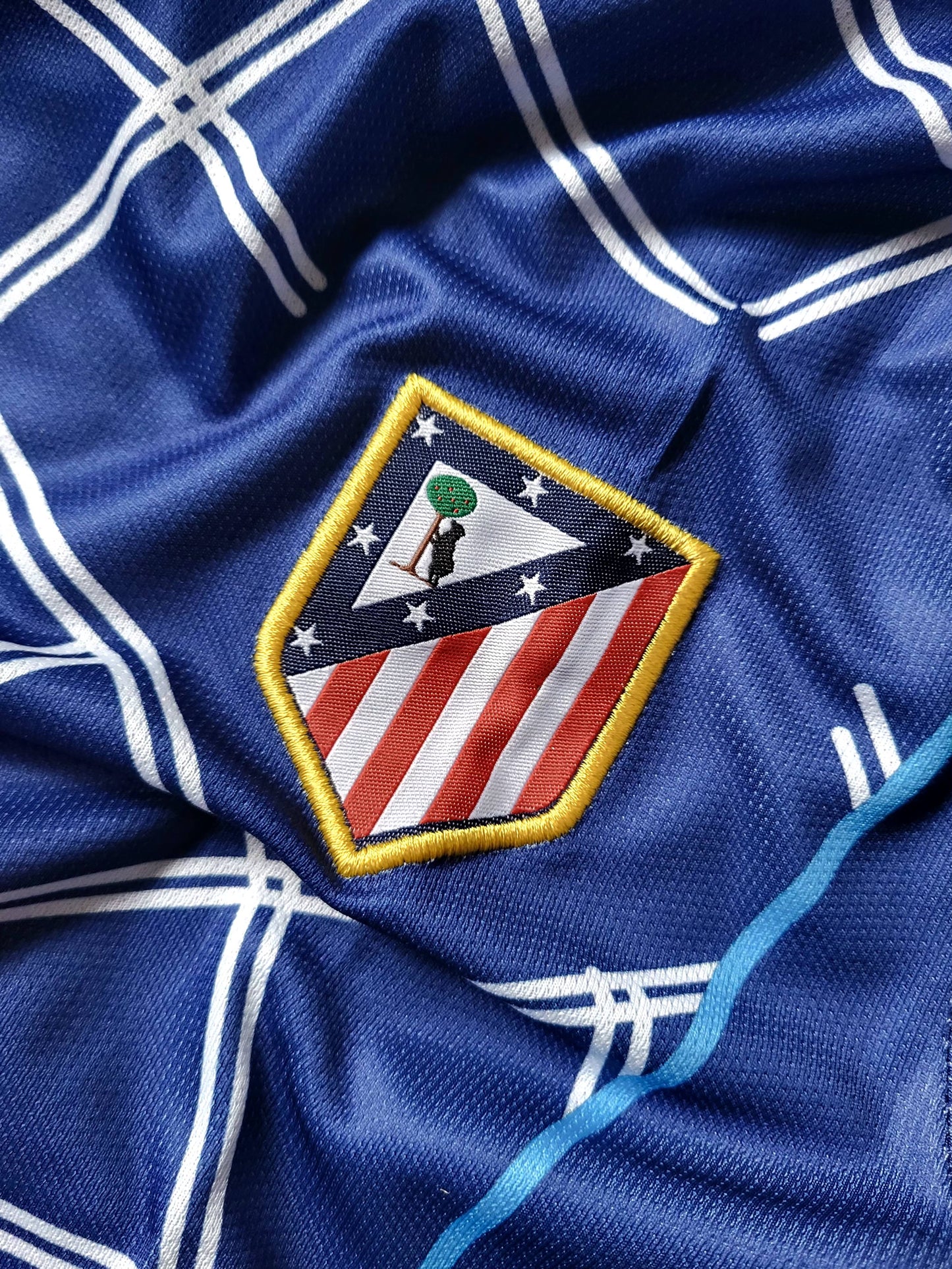 Close-up of a blue athletic jersey with white grid lines and an embroidered Atletico Madrid crest patch.