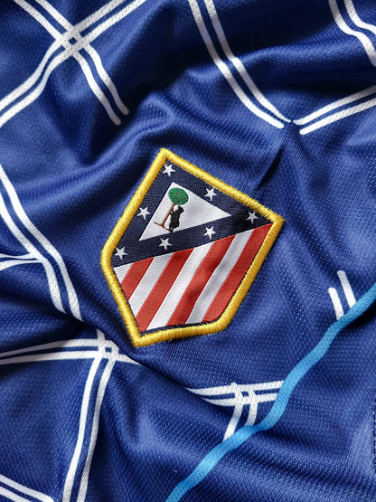 Close-up of a blue athletic jersey with white grid lines and an embroidered Atletico Madrid crest patch.
