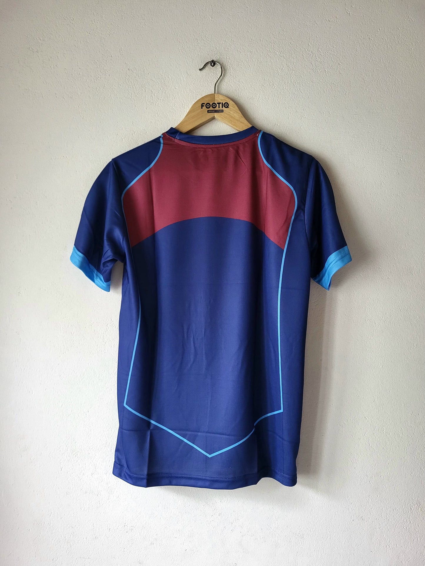 A short-sleeve athletic jersey hanging on a wooden hanger against a plain off-white wall. The back of the shirt is primarily dark blue with a large maroon block across the upper back, light blue piping lines, and light blue sleeve cuffs.