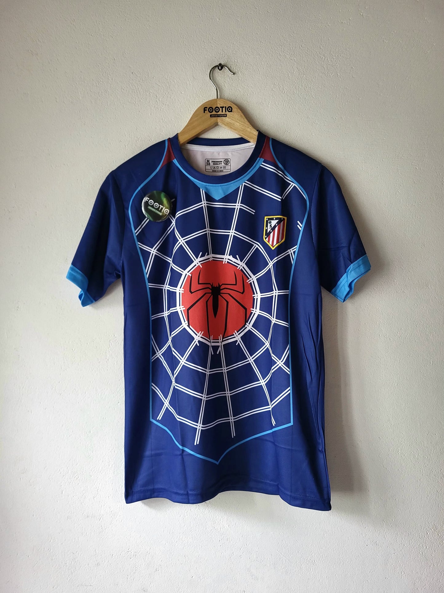 A blue short-sleeve Atletico Madrid soccer jersey with a Spider-Man web and red spider graphic on the front, hanging on a wooden hanger against a plain wall.