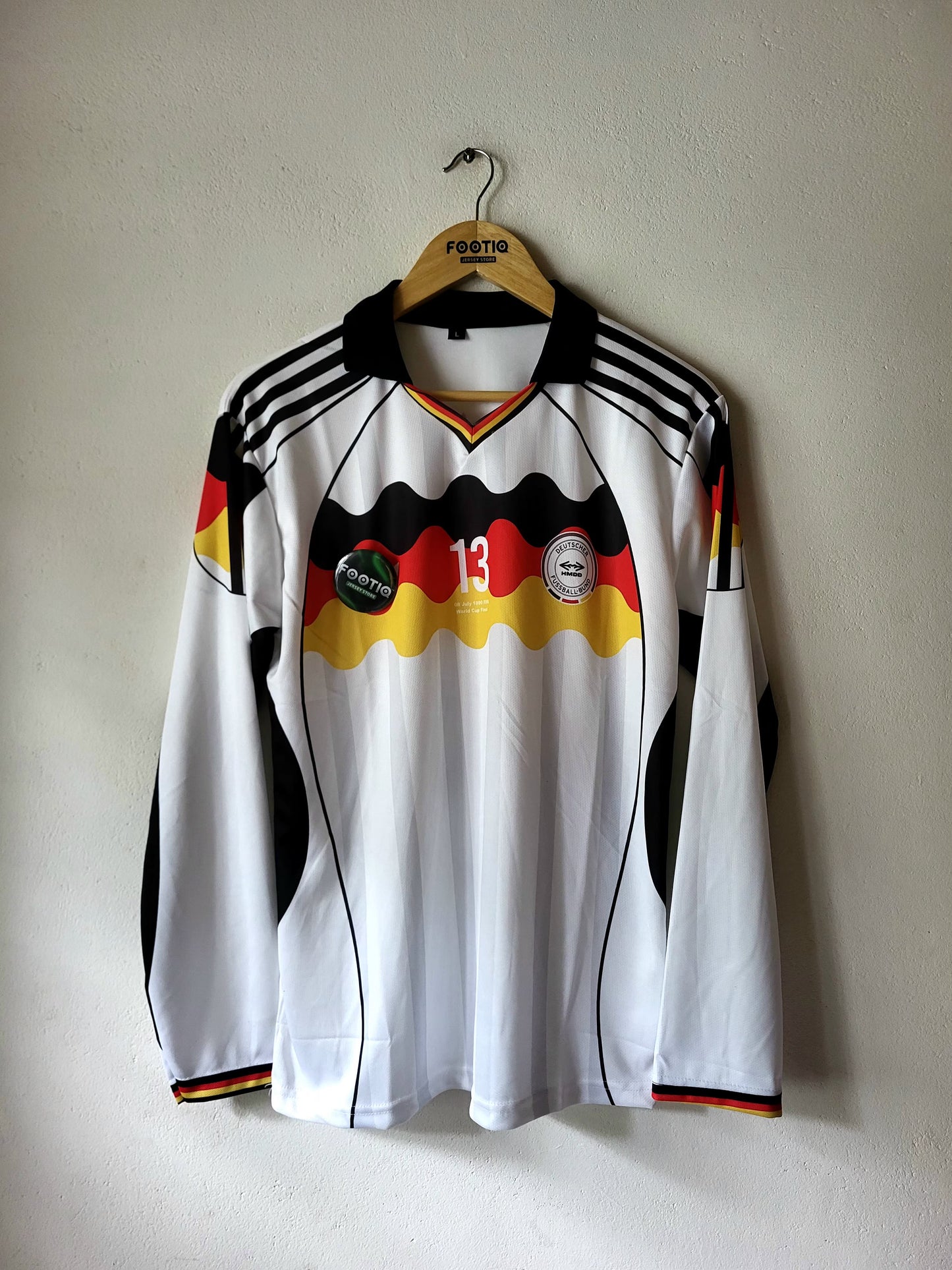 A long-sleeve white Germany national team soccer jersey with black and red accents, hanging on a wooden hanger against a plain wall.
