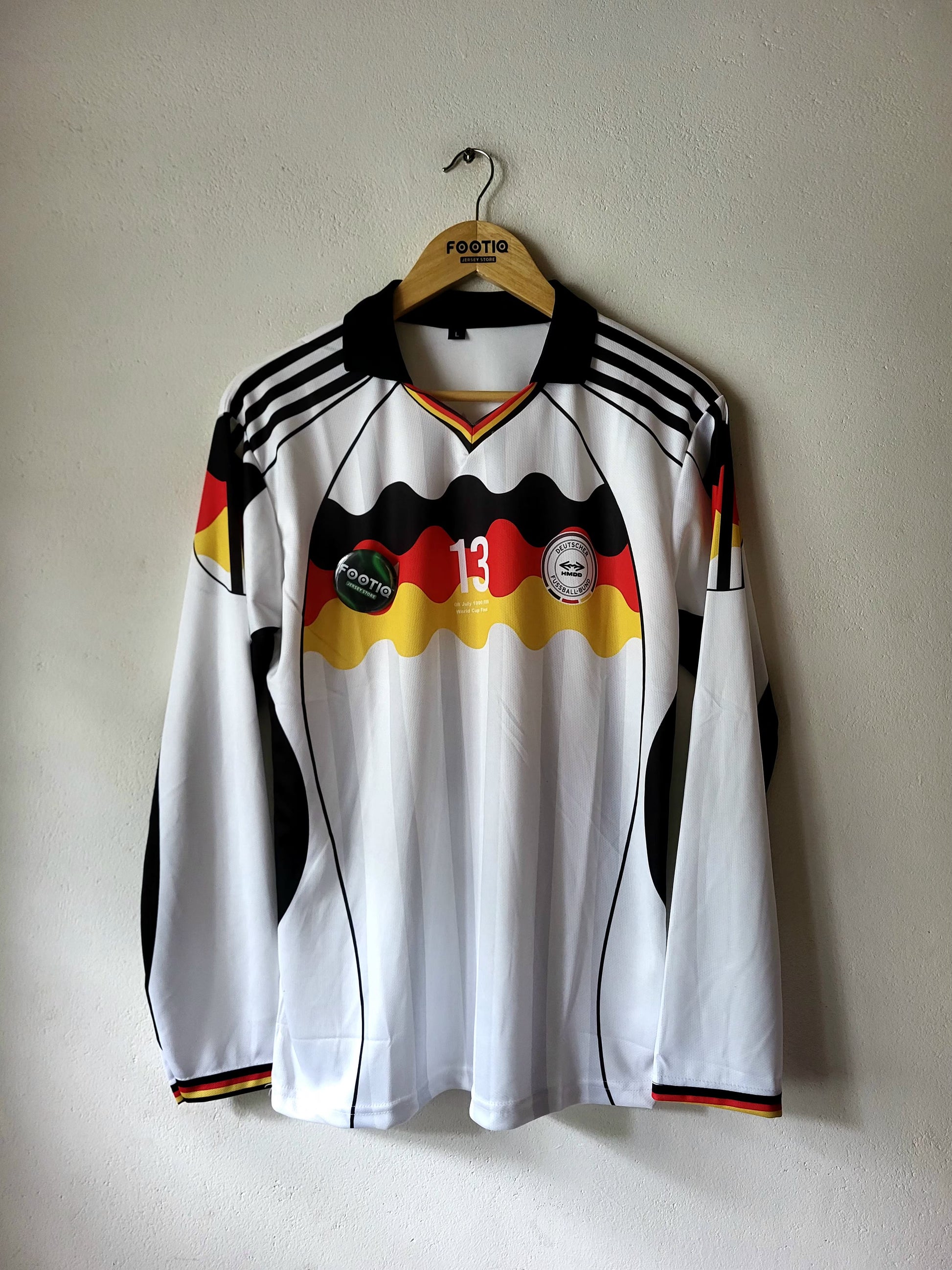 A long-sleeve white Germany national team soccer jersey with black and red accents, hanging on a wooden hanger against a plain wall.