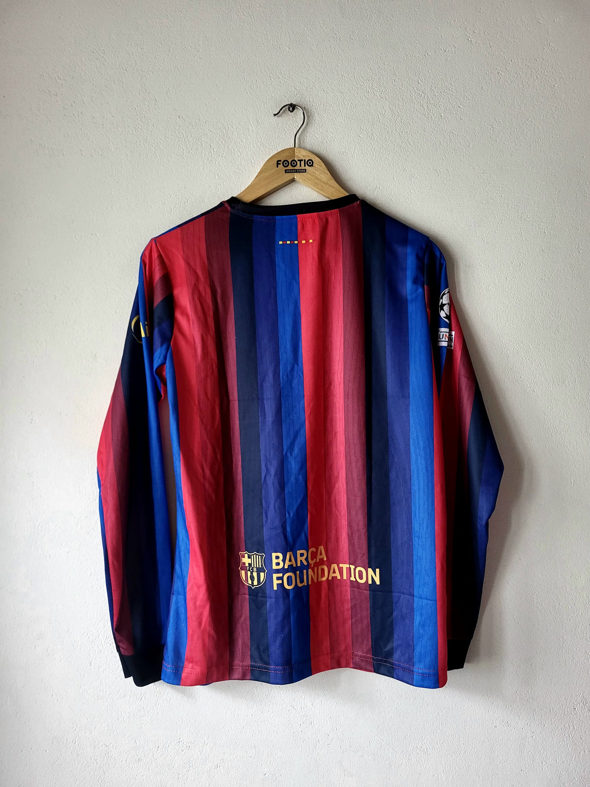 A vintage FC Barcelona long-sleeve soccer jersey with vertical red and blue stripes, hanging on a wooden hanger against a plain light gray wall. The jersey features gold 'BARCA' and 'VIP FOUNDATION' text on the front.