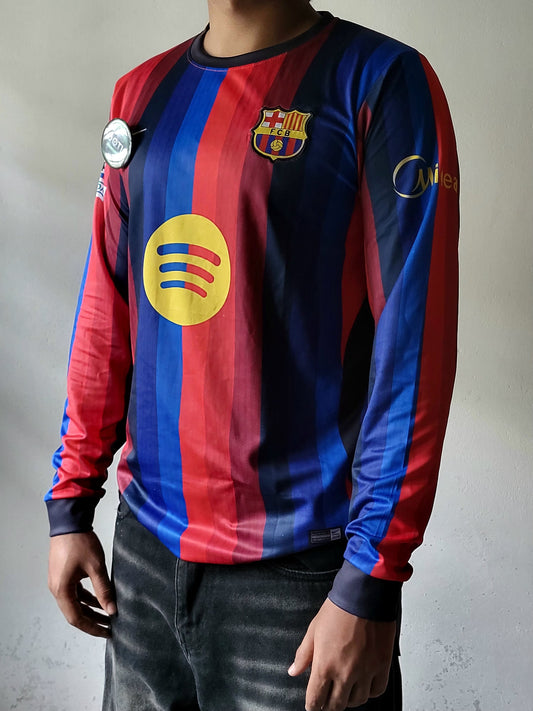 A person wearing a long-sleeve FC Barcelona soccer jersey with red and blue vertical stripes, a yellow Spotify sponsor logo on the chest, and the club crest on the left chest, paired with dark jeans against a neutral gray wall.