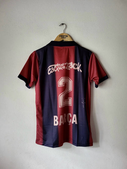A Barcelona soccer jersey with 'Cactus jack' and the number 2 on the back, hanging on a wooden hanger against a light gray wall.