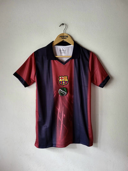 A vintage FC Barcelona soccer jersey hanging on a wooden hanger against a plain off-white wall. The jersey features a vertical color-blocked design with dark navy blue on the top half, a red-to-purple gradient in the middle, and dark navy on the bottom half.