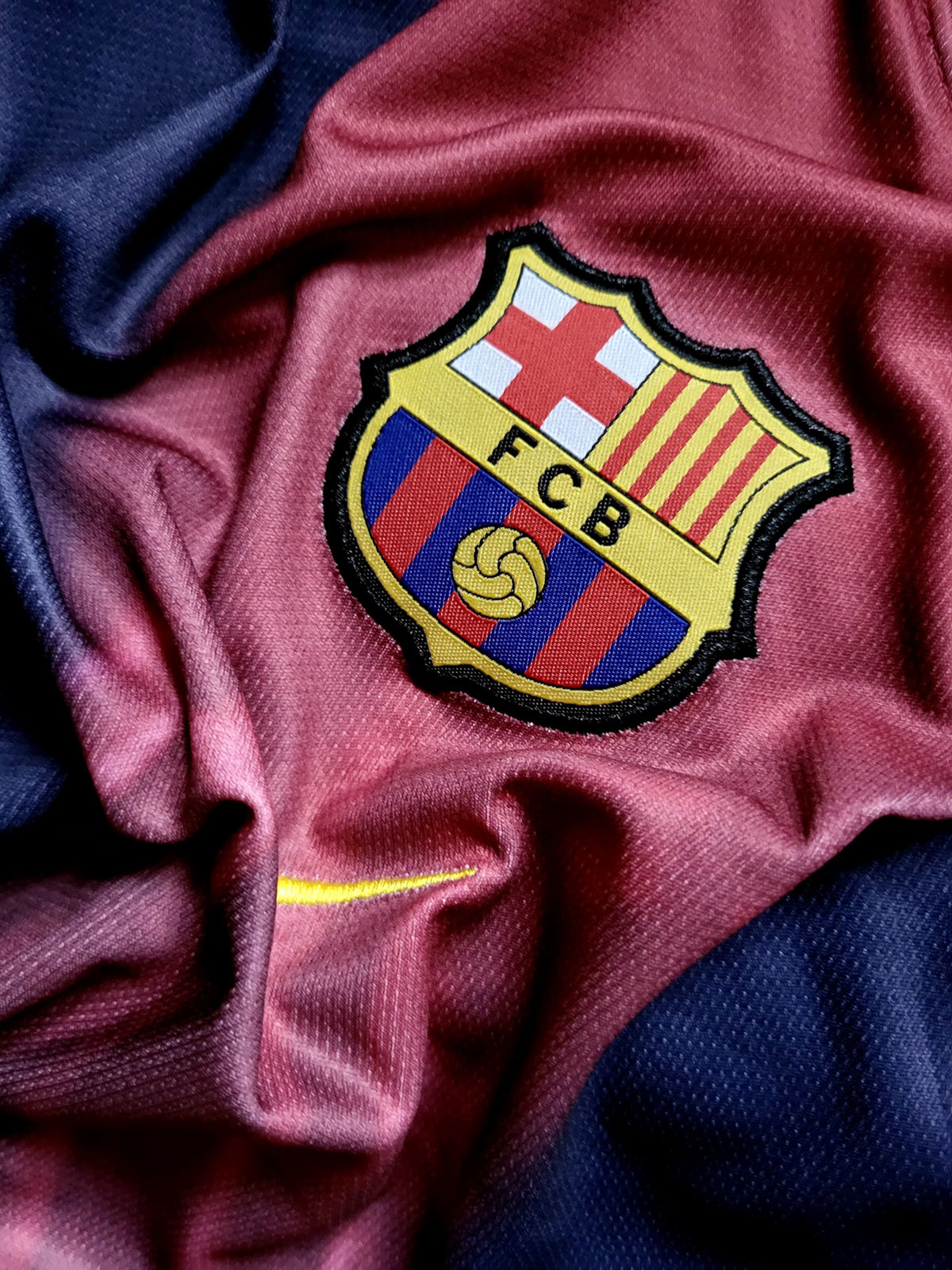 Close-up of an FC Barcelona soccer jersey fabric with the embroidered club crest centered on a draped maroon and navy background.