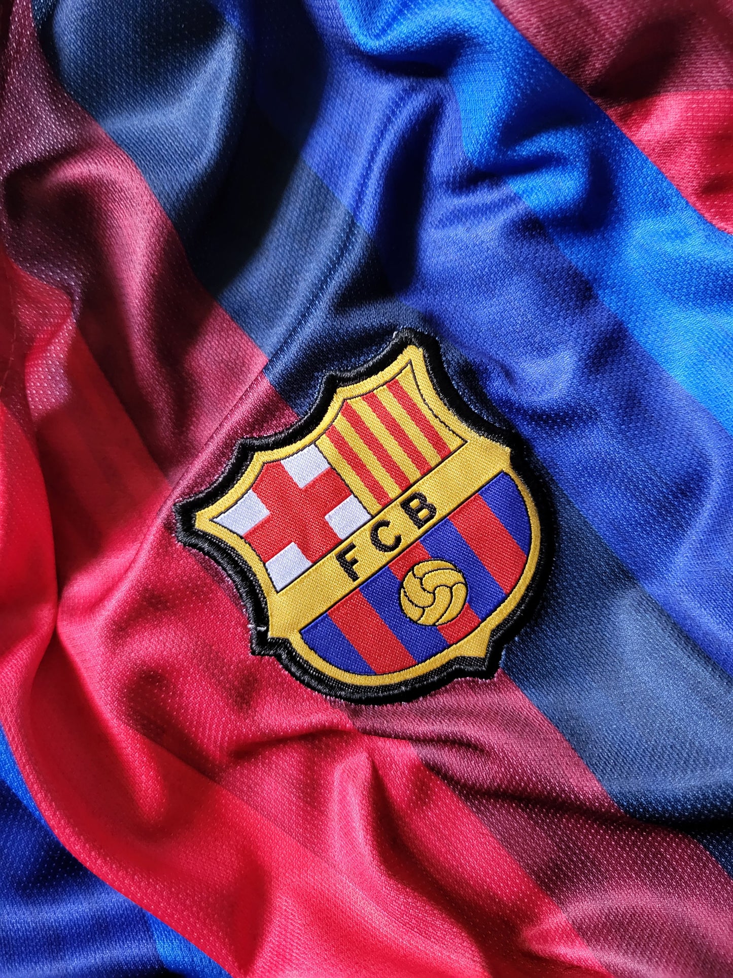 A close-up of a FC Barcelona soccer jersey fabric with the embroidered club crest centered on the chest. The jersey has the team's signature red and blue stripes with a satin-like sheen.