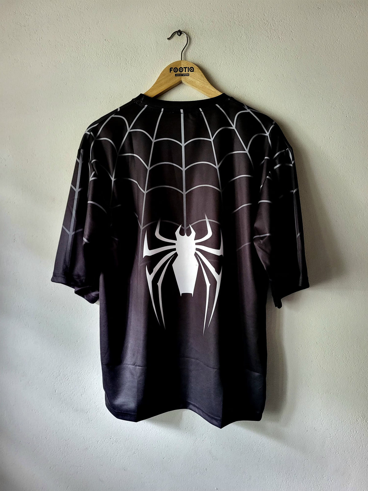 A black short-sleeve t-shirt with a white spiderweb pattern and a large white spider graphic on the front, hanging on a wooden hanger against a light gray wall.