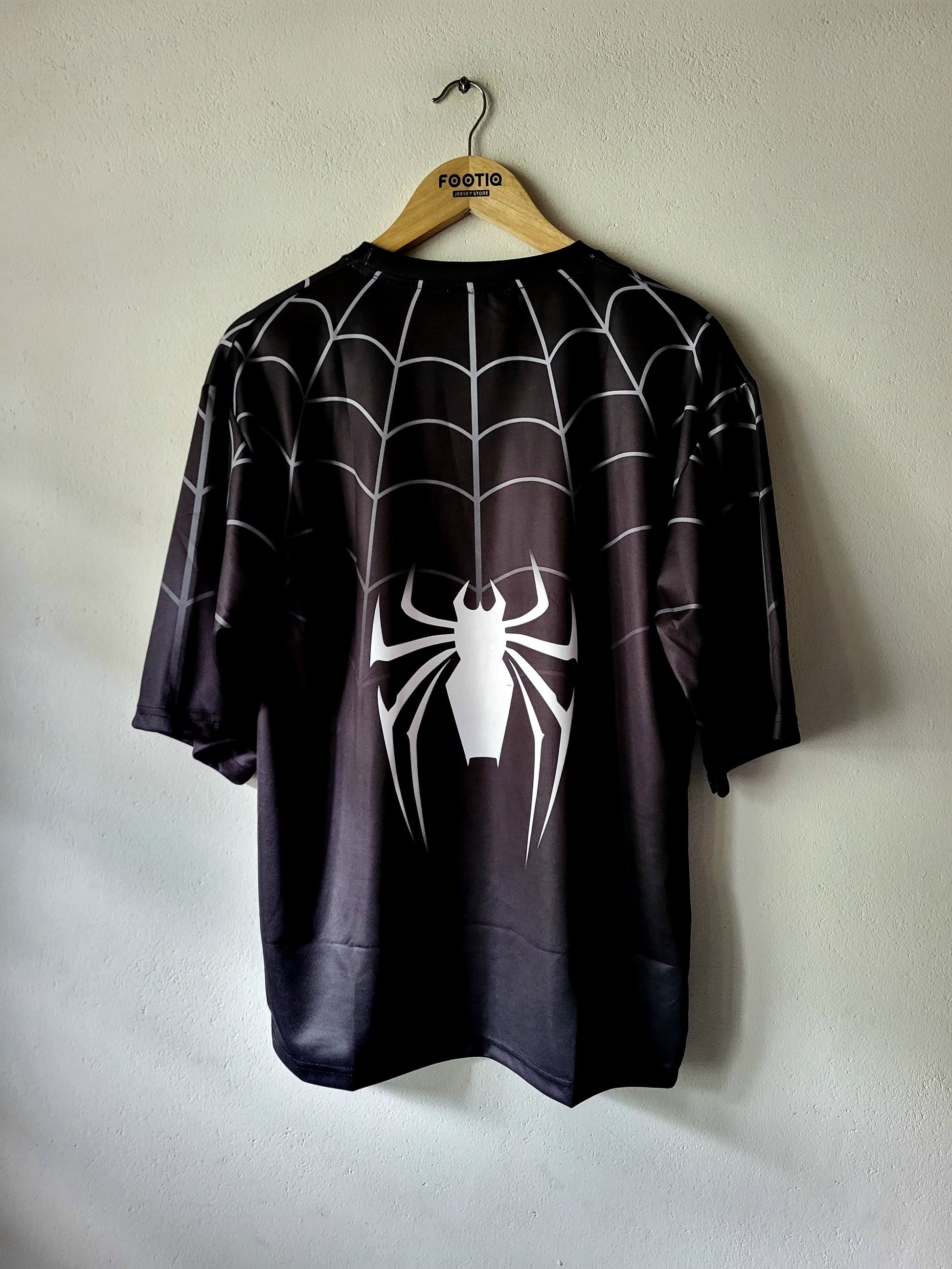 A black short-sleeve t-shirt with a white spiderweb pattern and a large white spider graphic on the front, hanging on a wooden hanger against a light gray wall.