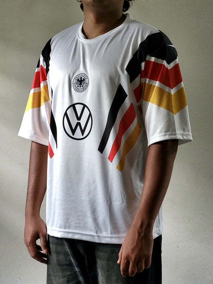 A person wearing a white Germany national team soccer jersey with black, red, and yellow sleeve stripes, featuring the Volkswagen logo on the chest.