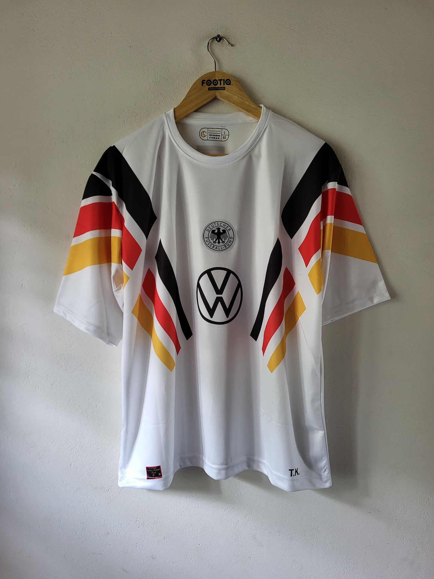 A white short-sleeve soccer jersey with black, red, and yellow diagonal stripes on the sleeves, featuring the Volkswagen logo and German flag emblem on the front, hanging on a wooden hanger against a plain wall.