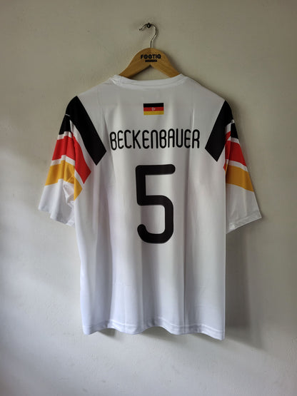A white Germany national team soccer jersey with 'BECKENBAUER' and the number '5' printed on the back, hanging on a wooden hanger against a plain wall.