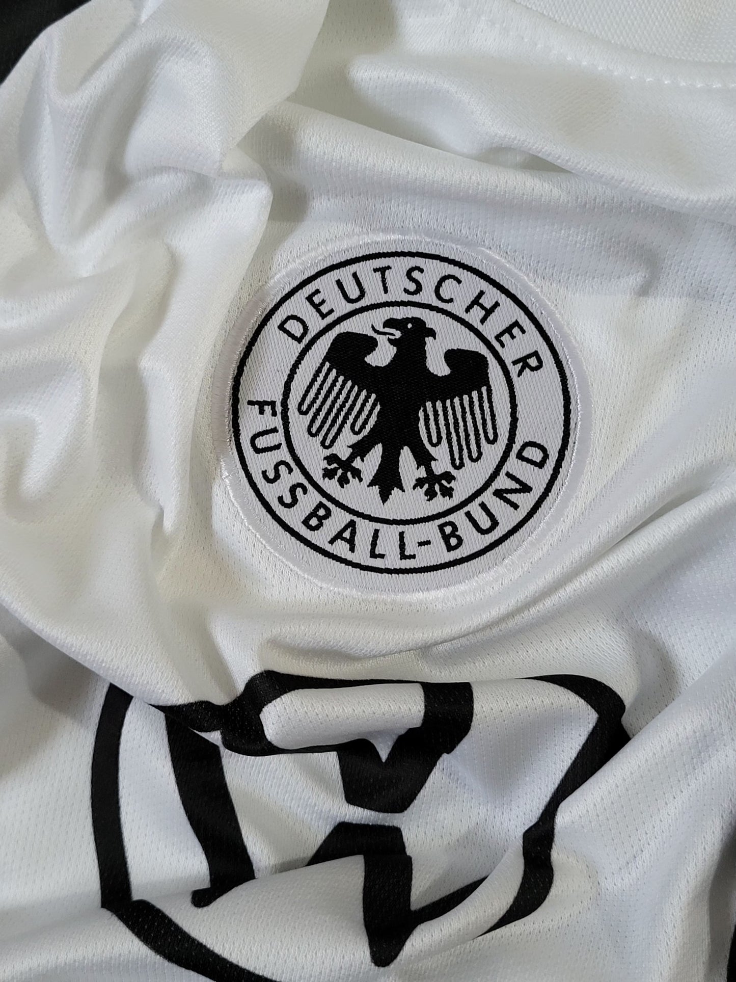 Close-up of a white soccer jersey with the circular black-and-white Deutscher Fussball-Bund crest on the chest and a large black logo below it.