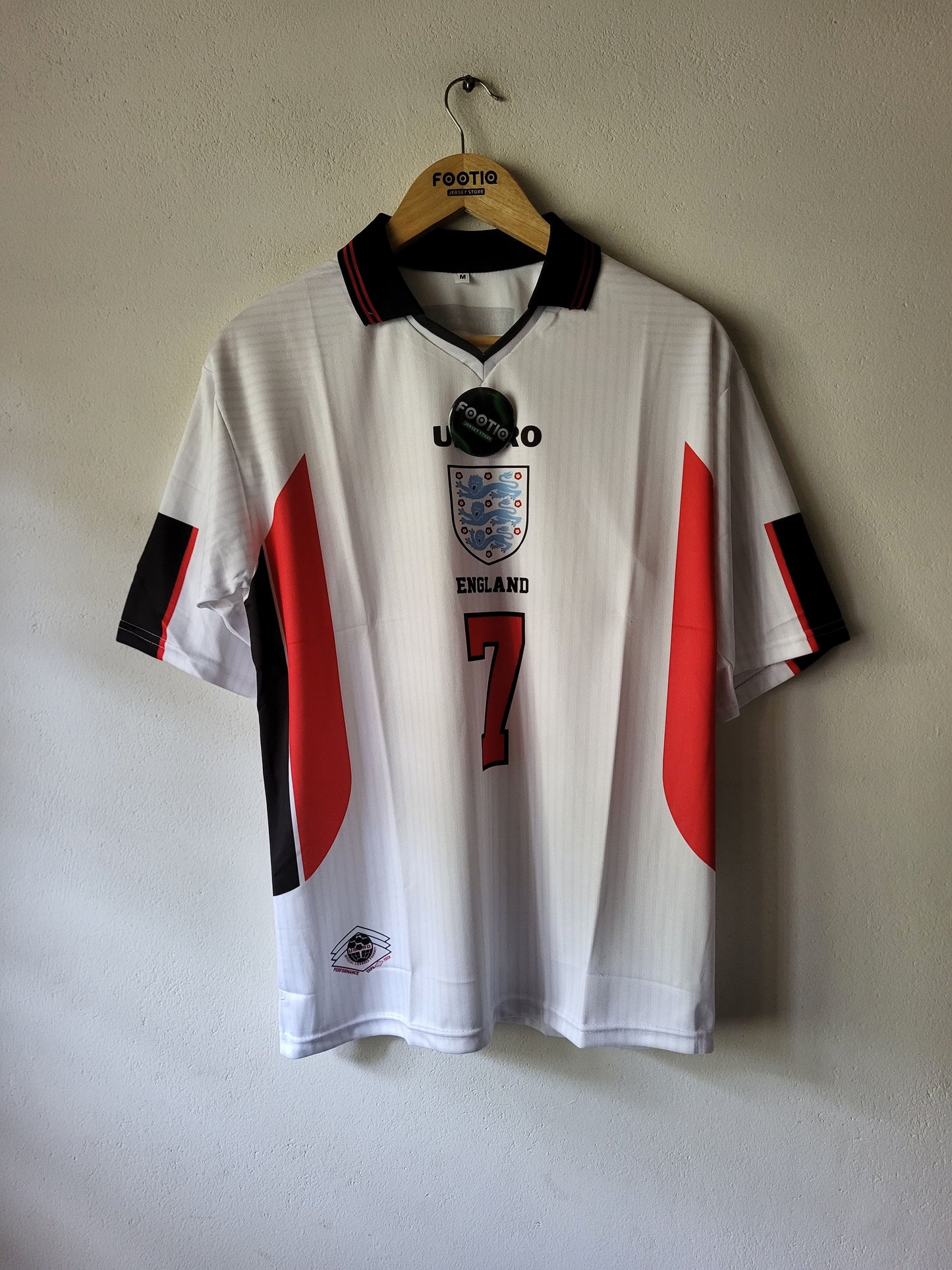 A white England national football team jersey with red and black side panels, hanging on a wooden hanger against a plain wall. The jersey features the England crest, the number 7 on the front, and a black hangtag attached near the collar.