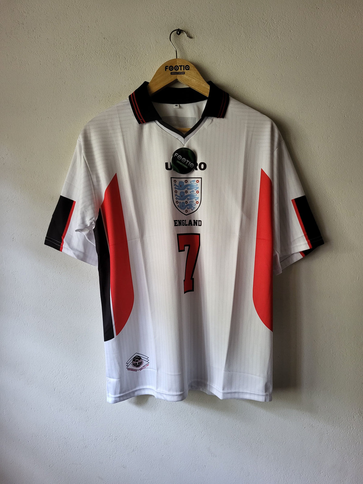A white England national football team jersey with red and black side panels, hanging on a wooden hanger against a plain wall. The jersey features the England crest, the number 7 on the front, and a black hangtag attached near the collar.