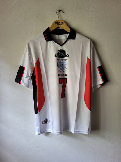 A white England national football team jersey with red and black side panels, hanging on a wooden hanger against a plain wall. The jersey features the England crest, the number 7 on the front, and a black hangtag attached near the collar.