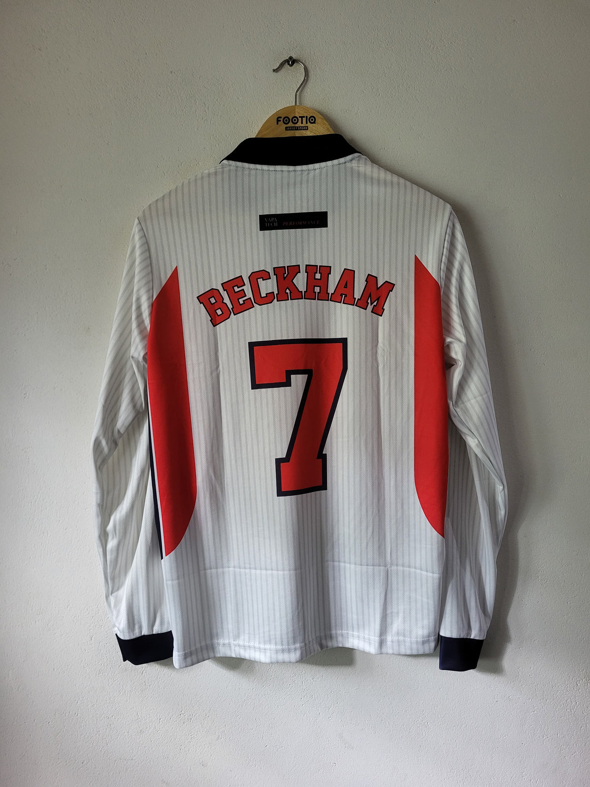 A white long-sleeve football jersey with 'BECKHAM' and the number '7' in red on the back, hanging on a wooden hanger against a plain wall.